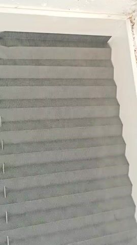 The video displays a close-up, shaky view of a grey pleated blind. The blind is partially obscuring a window, with the top edge of a white window frame visible at the very top of the first frame. Faint light suggests daylight is coming from behind the blind. The pleats of the blind are uniformly spaced and create a textured surface. In one frame, a small white object with a circular dial and some text is visible to the left of the blind. The lighting is muted, and the overall impression is of an indoor setting.