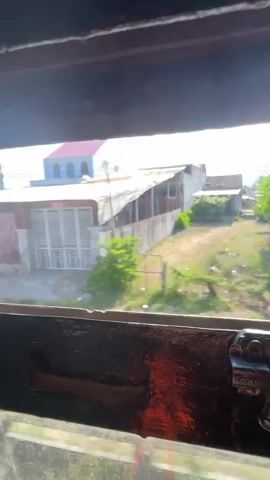 The video opens with a view through a window of a rural landscape with houses and trees. The camera then moves inside, showing a window with bars, then a doorway with blue doors, and finally the interior of a train carriage. There are people visible in the distance inside the train.
