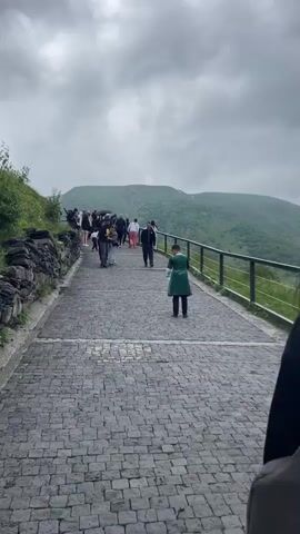 A group of people are gathered on a cobblestone path on a cloudy day, with a railing along the edge overlooking a lush green mountainous landscape. Some people are walking, while others are standing and looking out at the view. One person in the foreground is mostly obscured, but wears a backpack. Further down the path, a person in a green outfit stands looking out, and a group of people in casual attire are walking up the path. Towards the end of the video, a woman in a pink shirt holds up a Georgian flag. The mountains are covered in green vegetation, and the sky is filled with dramatic grey clouds.