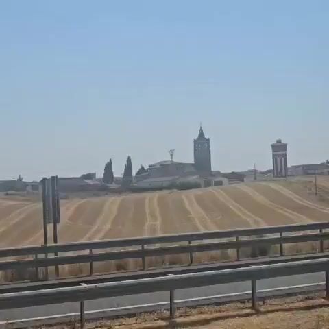 This video shows a rural landscape with a town in the distance. The foreground features a highway guardrail and asphalt. Beyond the highway, there is a harvested wheat field with circular tracks. In the background, a town is visible with a tall, dark church tower and a water tower. The sky is clear and blue. The video appears to be filmed from a moving vehicle, likely on the highway.