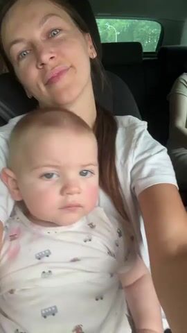 This is a selfie-style video taken inside a car, likely during the daytime given the natural light filtering into the vehicle. The primary subjects are a woman holding a baby, with a third person visible in the background. The woman appears to be speaking to the camera, exhibiting a range of emotions from smiles and laughter to more animated expressions. The baby, nestled in her arms, looks directly at the camera with a more neutral or slightly serious expression.

The woman is wearing a white t-shirt. The baby is wearing a onesie with a pattern of buses and cars. The background shows the interior of a car, including seats and headrests, and through the windows, glimpses of greenery suggest an outdoor setting, possibly rural or suburban. The presence of a car and the nature of the footage indicate travel or a journey. The woman's expressions and the way she interacts with the camera suggest a casual and personal recording, perhaps for sharing with friends or family. There is no visible text in the media file.