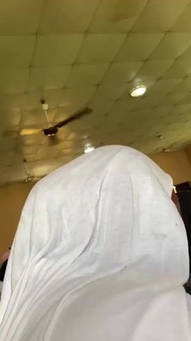 The video depicts a classroom setting with students seated at tables, most of whom are wearing head coverings. A person stands at the front near a whiteboard, seemingly lecturing or addressing the class. The room has a tiled ceiling, ceiling fans, and windows providing natural light. The overall atmosphere seems to be one of a formal learning environment.