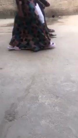 The video shows the lower part of a person wearing a long, patterned skirt and light purple sneakers, walking on a concrete surface. Another person, possibly a child, is walking alongside. There is a brick wall in the background and some plants.  The ground appears to be in an outdoor area. The camera angle is low. There is music playing in the background.