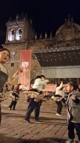 In a nighttime scene, multiple dancers, predominantly women, perform a spinning dance on a cobblestone plaza in front of a large, ornate stone building. The building, likely a cathedral, features a clock, spires, and illuminated windows. A banner with the word "Cusco" in bold letters stands prominently on a stage behind the dancers, which suggests they are dancing in Cusco, Peru. The stage is equipped with lighting fixtures and appears set for a performance. The dancers are wearing casual clothing, including jackets, sweaters, and pants, and are twirling their skirts, creating a dynamic visual effect. Several people watch the performance from nearby steps. The cobblestone plaza is illuminated by streetlights and lights emanating from the stage and the cathedral. The sky is dark, indicating the late hour of the performance.