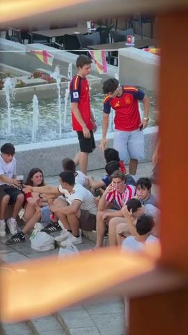 A group of young people are gathered around a fountain. Many are wearing red and yellow shirts, appearing to be supporters of a sports team. Two young men in red shirts with the number 17 and an emblem on the front are standing and talking. A group of others are sitting on the ground near the fountain. One young man is on his phone, and another is holding a can. The environment appears to be an outdoor public space, possibly a plaza, with tables and chairs visible in the background.