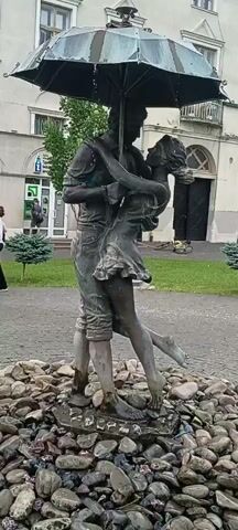 The video shows a bronze sculpture of a couple embracing under an umbrella. The sculpture is located outdoors in what appears to be a public space, possibly a park or plaza, with cobblestone pavement and some greenery in the background. There is a building with windows and a lamppost visible behind the sculpture. The umbrella appears to be made of metal and is tilted, with the couple leaning into each other as if dancing in the rain. The sculpture is surrounded by rocks and has a small water feature at its base.