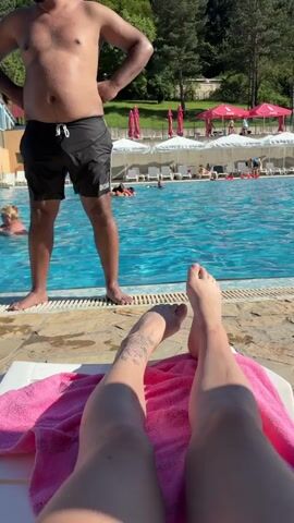 The video is an eye-level POV shot from a lounge chair, looking out over a swimming pool on a sunny day. In the foreground, a person's legs are visible, resting on a pink towel on a white lounge chair. A tattoo is visible on the person's left shin. To the left, a man wearing black swim trunks stands at the edge of the pool. In the background, the swimming pool is filled with people swimming and playing. Along the far side of the pool are rows of white lounge chairs with umbrellas and a wooded hillside. A yellow and red water slide is visible in the distance.