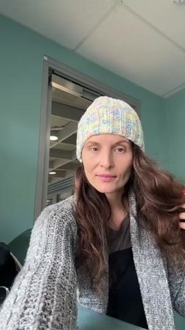 A woman with long, brown hair and a knitted hat is shown in a room. She is wearing a gray cardigan over a black top. The woman is touching her hair and looking at the camera. There is a glass door in the background, and a desk with glasses in the foreground. The room appears to be an office or a waiting area.
