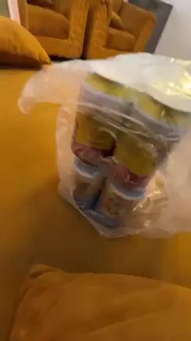 A plastic bag containing several cans of food or formula sits on a mustard-colored couch or sofa. The couches have a ridged or textured pattern. The plastic bag obscures the labels on the cans, but the colors indicate a mix of products.  The scene is indoors with neutral lighting. The camera is positioned at an angle looking slightly down at the bag on the couch.