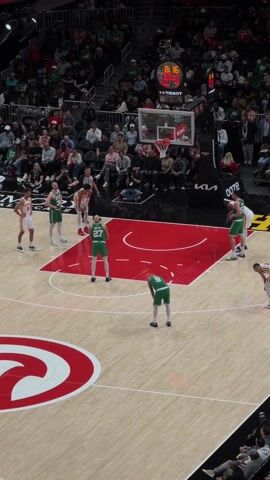 The video depicts a basketball game in progress. The court is red and light brown with markings and logos. The players wear green and white uniforms or red and white uniforms. A player in green and white is shooting a free throw, while other players line up along the edge of the free throw lane, awaiting the rebound. Behind them, a packed audience sits in the stands. A scoreboard is visible above the court.