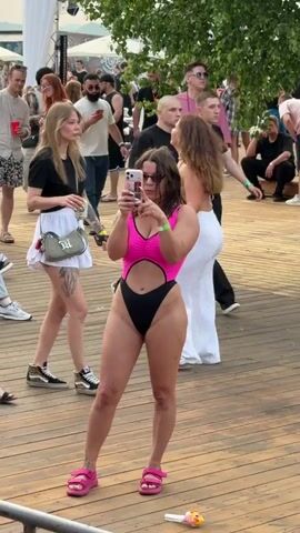 The video shows a group of people at an outdoor event, likely a festival or concert. The main focus is a woman in a pink and black one-piece swimsuit, holding up her phone to take a picture or video. She is standing on a wooden deck, and other people are visible in the background. Some are dressed casually, while others are in swimwear. The atmosphere appears to be lively and social.