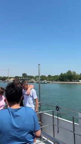 The video shows a group of people on a boat, enjoying a sunny day on the water. The boat is moving, as evidenced by the wake behind it.  The sky is blue with a few clouds. Some people are standing near the railing, looking out at the water and coastline.  A yellow crane is visible in the distance, as well as some docks and vegetation. The boat's white roof and support structures are also visible.
