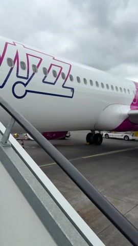 The video shows a view from a set of stairs looking at the side of a Wizz Air passenger plane with pink and white livery. The camera pans to the right to show the back of the Wizz Air plane, including its wing and engine. Beyond the airplane, there are people walking on the tarmac, a hillside with buildings, and another set of stairs leading up to the back of the plane. The camera continues panning to the right to show a Ryanair plane in the background, surrounded by cones. The sky is overcast.