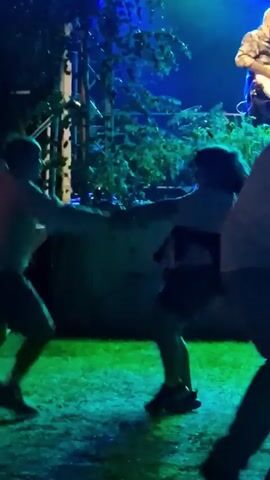 A dimly lit outdoor scene with green and blue lighting effects. In the foreground, two blurred figures are dancing. The figure on the left appears to be wearing a light-colored top and dark shorts, while the figure on the right is wearing a dark top and skirt. They are facing each other and their bodies are in motion, suggesting they are actively dancing. In the background, on a raised platform, a bald man in a patterned shirt is playing an electric guitar. He is illuminated by a bright blue light. Lush green foliage is visible to the left and behind the guitarist, partially obscuring the stage structure. A portion of a railing or fence is visible on the far left of the frame. The ground appears to be grass, covered in patches of green light. The overall atmosphere suggests a live music performance outdoors at night.