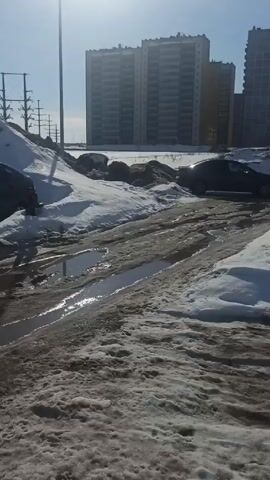 The video shows a muddy, slushy parking lot on a sunny day. There is a large puddle of water in the center of the lot, with tire tracks visible through the mud and slush. Several cars are parked in the lot, some covered in dirt. In the background, there is a large building and a pile of snow. A white van drives through the puddle, splashing water. A tow rope is visible laying in the puddle. The camera pans to show more cars parked in the lot.