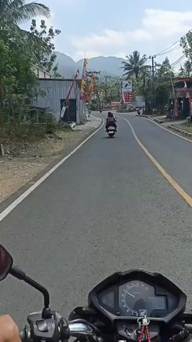The video is a first-person view from a motorcycle, showing a road with a few other vehicles and buildings. In the foreground, the motorcycle's handlebars and speedometer are visible. Ahead, a person on a scooter is riding in the same direction. To the left of the road are some buildings, one with a large orange banner. To the right, there's a gas station with prominent red and white signage, and more buildings. In the background, mountains and trees are visible under a clear sky. The road appears to be paved and has yellow lane markings. The overall impression is a sunny day in a rural or semi-rural area.