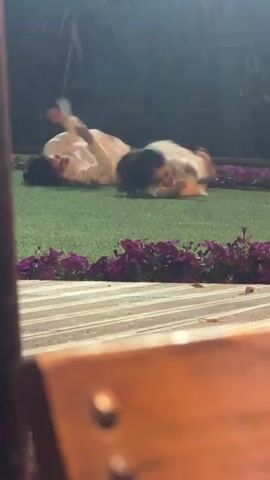 The video shows two children rolling and flailing on a patch of artificial grass at night. They are both dressed in light-colored shirts and dark pants. Purple flowers line the edge of the grass. The view is partially obstructed by wooden furniture.