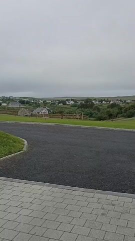 The video shows a grey, overcast sky over a rural landscape. In the foreground, there is a paved driveway with a cobblestone border and a patch of green grass. Beyond the driveway, there is a field with a wooden fence, a playground set with swings, and several houses scattered across a gentle slope. The houses are white with darker roofs and appear to be part of a village or small town. The overall scene is quiet and appears to be a still, uneventful moment in time.