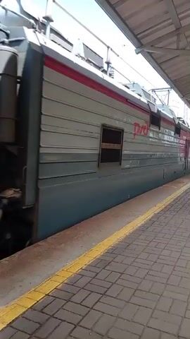 A train is moving past a stationary camera on a train platform. The train is composed of an electric locomotive and several tank cars. The locomotive is red and gray with the logo RZD on its side. The tank cars are various colors, including brown, green, and red. One tank car has the text HHK on its side. The platform is paved with brick and has a yellow line along the edge. The sky is blue and clear, and there are trees visible in the background. The camera appears to be mounted at eye level.