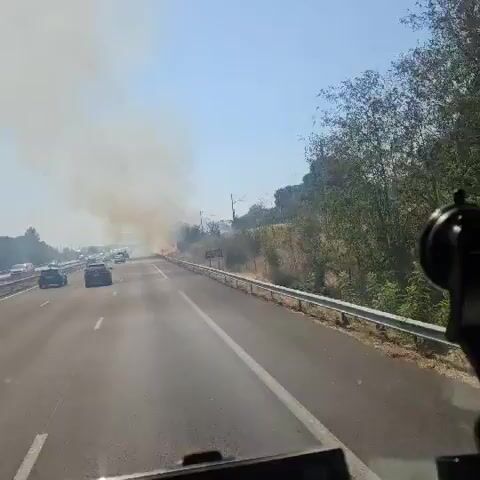 A video captured from the perspective of a vehicle driving on a highway shows a roadside fire. Thick smoke billows into the clear blue sky from the right side of the road. Several cars and a white van are visible on the highway ahead, with some appearing to be slowed down or stopped due to the fire. The fire itself is visible as a line of flames near the edge of the road, interspersed with trees and dry vegetation. A metal guardrail separates the highway lanes from the embankment where the fire is burning. Overhead power lines are visible on the embankment. The interior of the vehicle shows a dashboard with a GPS device and a camera mount. The sun is shining brightly.