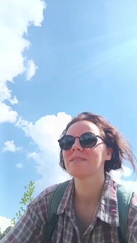 A medium shot, low-angle selfie-style video shows a person with short brown hair wearing sunglasses and a plaid shirt. They have a backpack on. The person is looking slightly off-camera to their right. The background is a bright blue sky with scattered white clouds. The video appears to be filmed outdoors on a sunny day.
