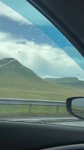 The video is shot from the inside of a vehicle, looking out the side window. The window has raindrops on it. In the distance, there are rolling green hills with some patches of white, possibly snow or rocks. The sky is cloudy with patches of blue. A silver car passes by on the road in the middle ground. A metal guardrail runs along the side of the road.