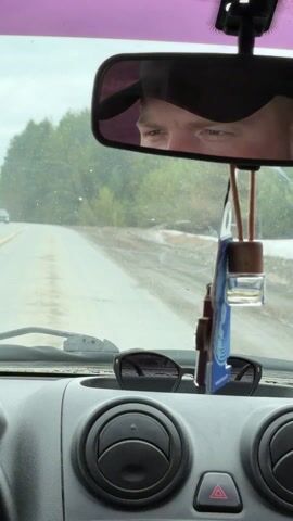 The video shows a view from the inside of a car driving on a slightly worn road. A sparse forest lines the roadside. Through the windshield, another vehicle can be seen driving in the distance. A driver is visible in the rearview mirror. The car's dashboard is gray, with sunglasses placed in the center console. A car freshener hangs from the rearview mirror. The dashboard also contains a speedometer and circular air vents. The weather appears overcast.