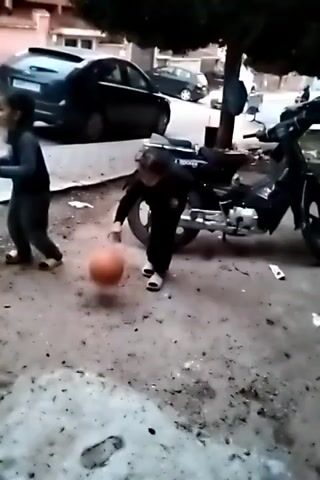 Outside on a cloudy day, a young child in a black outfit bounces an orange basketball on a grey surface that has scattered debris.  There are two dark cars parked on a curb next to the child.  There is also a black moped parked to the right of the child.  Another child is partially visible on the left of the screen.  A tree is planted in the background.