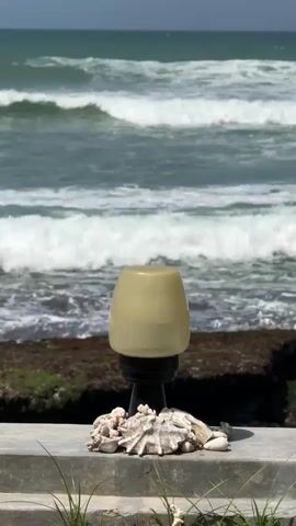 A vertical video shows a decorative lamp with shells at its base on a concrete ledge. Behind the lamp, the ocean waves crash against a rocky shore. The sky is a pale blue, and the overall scene is bright and sunny.