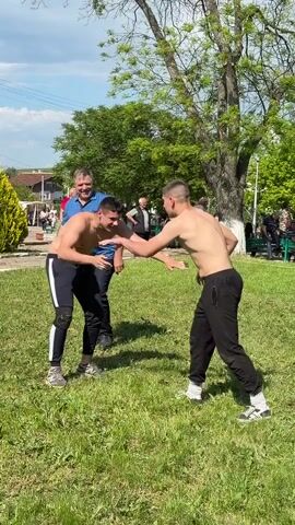 Two men are wrestling on a green lawn, surrounded by a small crowd of onlookers. The wrestlers are shirtless and wearing dark athletic pants and sneakers. A referee, dressed in a blue polo shirt and dark pants, watches closely. The wrestling takes place on a sunny day in what appears to be a village square, with trees, buildings, and a power pole in the background.