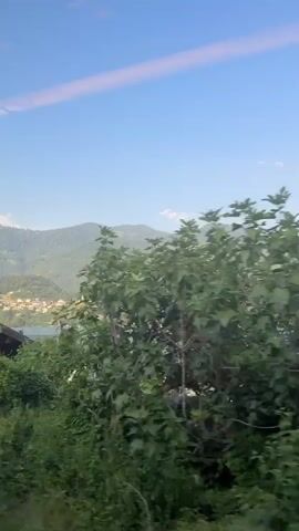The video shows a view from a moving vehicle, likely a train, looking out a window. The scene transitions from rural to more populated areas. Initially, there are glimpses of houses with dark roofs surrounded by greenery, with misty mountains in the background. As the vehicle moves, the view changes to a multi-story white building with a sign reading "ALBERGO PIZZERIA" in yellow letters. There are several windows visible, some with blue shutters. Further along, a man is seen standing near a parked car and a bicycle is visible. Trees and more mountains continue to feature in the landscape. The sky is a clear blue with some light streaks. The lighting suggests it is daytime, likely late afternoon or early evening.