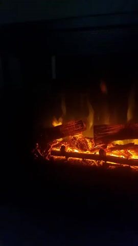 This is a close-up, low-light shot of an electric fireplace insert. Realistic-looking artificial logs are arranged on a bed of glowing orange embers. Subtle yellow and orange flames flicker behind and between the logs, creating the illusion of a real fire. The background is entirely black, emphasizing the warm glow of the fireplace. The video appears to be a short loop, showing the flames and embers moving slightly.