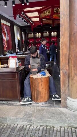 A man wearing glasses and an apron is sitting on a wooden stool outside a shop. He is working on a ring with a hammer.  The shop is decorated in a traditional Chinese style.  There are several people inside the shop, including two women in blue dresses who are watching the man work.