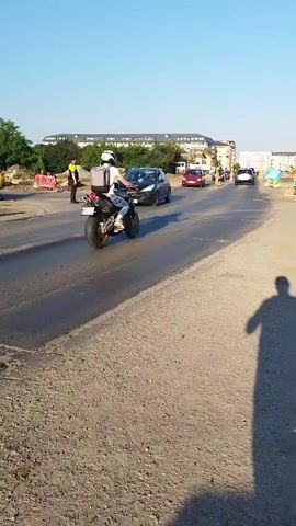 The video captures a street scene with ongoing construction. A motorcycle rider is seen in the foreground, followed by several cars. A person in a yellow vest, likely a construction worker, stands on the side of the road directing traffic. The road surface appears to be newly paved or undergoing work, with some areas of loose gravel and dirt. Buildings and trees are visible in the background under a clear blue sky. The overall impression is a busy street with traffic management in place due to construction activities.