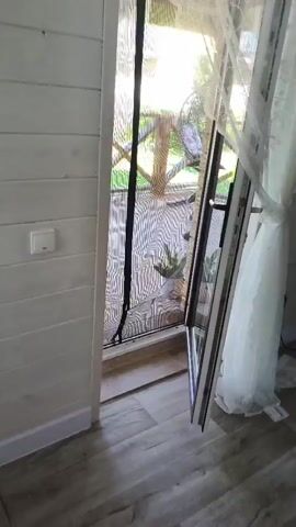 The video begins by showing a white wooden wall with a light switch. A doorway with a screen door is visible, and beyond that, a covered outdoor area with wooden flooring and a railing. There is a potted plant on the deck. The screen door appears to be made of a mesh material and has a black strip down the middle. As the video progresses, the camera moves through the screen door, revealing a green lawn and trees. The outdoor area has a thatched roof and curtains hanging from the railing. The video ends with a view of the deck and the surrounding greenery.