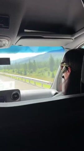A person wearing sunglasses and a cap is driving a vehicle. The view is from inside the vehicle, looking out the windshield. The road is visible, with a guardrail to the right. Beyond the road, there are lush green trees, hills, and mountains under a cloudy sky. A speed limit sign indicating 70 is visible by the side of the road. The person is looking forward, seemingly focused on the road.