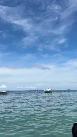 A white and green boat with several people on board sails across a calm blue sea under a cloudy sky. In the distance, a hazy landmass is visible on the horizon. The water in the foreground is clear and turquoise.