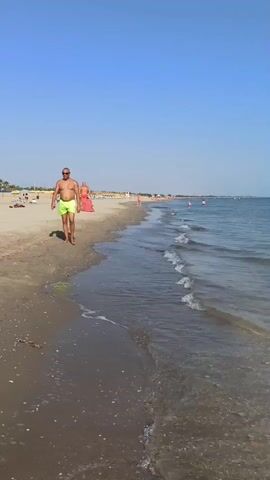The video shows a beach scene. In the foreground, a man wearing yellow swim trunks walks along the wet sand near the water's edge. His back is to the camera as he walks away from the viewer. To his left, further up the beach, are beach chairs and umbrellas, with other people visible in the distance. The sea is calm with small waves lapping the shore. The sky is clear and blue. The video pans slightly to the right, showing more of the sea and the distant beach.