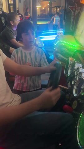 The video shows a boy in a white shirt playing a racing game in an arcade. The boy is seated and gripping the steering wheel, and there is another young boy in a tie-dye shirt standing beside him, smiling and laughing, with his hands in front of his chest. Several other people are visible in the background, including a woman behind the boy in the tie-dye shirt, and other figures standing near lit-up arcade machines. The arcade appears to be well-lit, with a glass wall and door visible in the background, showing an outdoor area with lights on. The arcade machines are brightly illuminated with various colors, including blue and red.