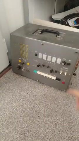 A hand is seen interacting with a vintage electronic device. The device is gray and box-shaped with a black handle on top. The front panel is filled with buttons, switches, and what appear to be indicator lights. The labels on the panel are in Cyrillic script. Visible text includes "ЦИК," "ГРАСЕД 1," "КАНАЛЫ," "СЕТЬ," "ЛИТ," "ПР-СЕТ," "КОНТУР ГРС," "МИР," "ГРС," "ГРА," and numbers 1 through 5 above corresponding buttons. The hand is pressing a button labeled "5" and then turning a knob labeled "СЕТЬ." The device is placed on a speckled gray carpet. The background shows a white wall and a glimpse of a doorway. The scene appears to be indoors, possibly in a storage or workshop area. The lighting is neutral, suggesting it is daytime or artificial lighting is in use. The overall impression is of a user operating an older piece of technology.