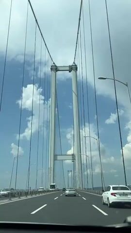 The video depicts a drive across a long suspension bridge on a clear day with a blue sky dotted with puffy white clouds. The bridge has two tall, white towers, each connected by thick cables extending down to the road surface. Several cars and a bus are visible on the multiple lanes of the bridge, with lampposts lining both sides of the road. The video is taken from inside a vehicle, looking forward towards the distant tower of the bridge.