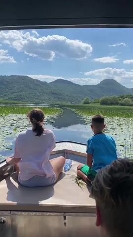 Two individuals, a woman and a boy, are seated in the front of a boat, facing away from the camera. They are looking out at a scenic lake surrounded by lush green mountains. The water is covered in lily pads, creating a textured surface. The sky is a vibrant blue with fluffy white clouds, suggesting a pleasant, sunny day.

The woman, with her hair tied back in a ponytail, wears a light-colored t-shirt and shorts. The boy, to her right, is wearing a blue t-shirt and green shorts. A plastic water bottle is placed between them on the boat's seating. In the foreground, partially visible, is the back of another person's head, indicating there are at least three people on the boat.

The boat appears to be moving, as suggested by the slight blur in the background and the perspective of the water. The overall mood is one of peaceful exploration and enjoyment of nature. The mountainous landscape and the vastness of the lake point to a natural, perhaps remote, location. The time of day appears to be daytime, likely mid-morning or afternoon, given the brightness of the sunlight and the clear sky.

There is no visible text in the media file. The scene captures a serene moment of a boat trip on a beautiful lake in a mountainous region.