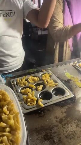 The video shows a person wearing a white t-shirt with "FENDI" printed on it, preparing food. The person is using a blue scoop to transfer macaroni from a clear plastic container into a metal mold with circular compartments. The mold is placed on a dark, worn surface, likely a griddle or food preparation station. Another person, wearing a purple hijab and a gold-patterned dress, is visible in the background, holding a small plastic bag with white contents. The environment appears to be outdoors or in a food stall setting, with dim lighting and some steam or smoke in the background. The focus is on the preparation of the macaroni, which is arranged in the compartments of the mold.