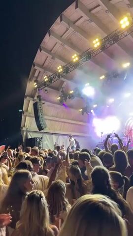 The video captures a live concert at night, viewed from within a dense crowd. The stage is brightly lit with pink and purple lights, and spotlights illuminate the performers. A large speaker hangs from the scaffolding above the stage. Many people in the audience are holding up their phones to record or photograph the event, with one phone prominently displaying the camera interface. The overall atmosphere is energetic, with people appearing to dance and enjoy the music. The background features trees and the night sky.