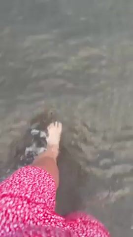 A first-person point-of-view shot shows a person wading through shallow water. The person is wearing a red and white patterned garment, likely shorts or a skirt. The water is murky and the bottom is visible. The person's bare feet are splashing in the water as they walk.