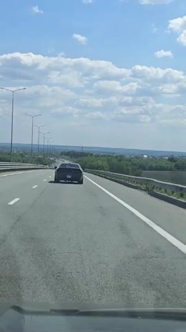 A driver films the rear of a black Dodge Challenger as they travel down a highway on a clear, sunny day. The Challenger is in the center lane of the three-lane highway. The road curves gently to the left, with a guardrail and green trees visible on the right. Streetlights line the highway to the left. Other cars can be seen in the distance. White lane markings are visible. The sky is blue with scattered white clouds. The driver is using a dashboard camera, and the interior of their car is visible at the bottom of the frame. Music is playing in the background.