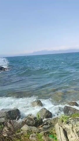 The video shows a choppy blue ocean with waves crashing against a rocky shore under a clear blue sky. The waves are white and foamy as they hit the rocks. The rocks are large and varied in shape and size, with some patches of green vegetation growing between them. In the distance, there is a hazy line of what appears to be mountains or clouds on the horizon. The overall impression is of a natural, somewhat wild coastal environment.