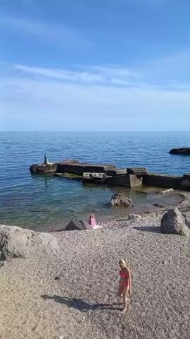 The video shows a scenic view of a beach on a sunny day. The camera pans across the scene, revealing a rocky coastline with clear blue water. In the foreground, a person in a pink bikini walks along the pebble beach. Further back, a person in a blue bikini is visible near the water's edge. On a rocky outcrop in the sea, a small statue stands. The background features a clear blue sky with scattered clouds and a coastline with trees and buildings. The video captures a peaceful and idyllic beach environment.