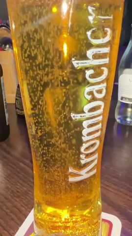 A close-up shot of a tall glass of Krombacher beer. The beer is golden yellow and filled with rising bubbles. The glass has the Krombacher logo printed vertically on it in white lettering. The glass sits on a coaster on a wooden surface. In the background, out of focus, there are bottles and other bar-like elements.