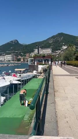 The video shows a man, wearing shorts and no shirt, standing on a green carpeted surface next to several boats docked in a harbor. He is bent over and appears to be working with some items on the ground. Behind him, a walkway with a railing separates the dock area from a path where several people are walking in the distance. The background features a body of water, buildings along the coast, and a large, green, mountainous landscape under a clear blue sky.